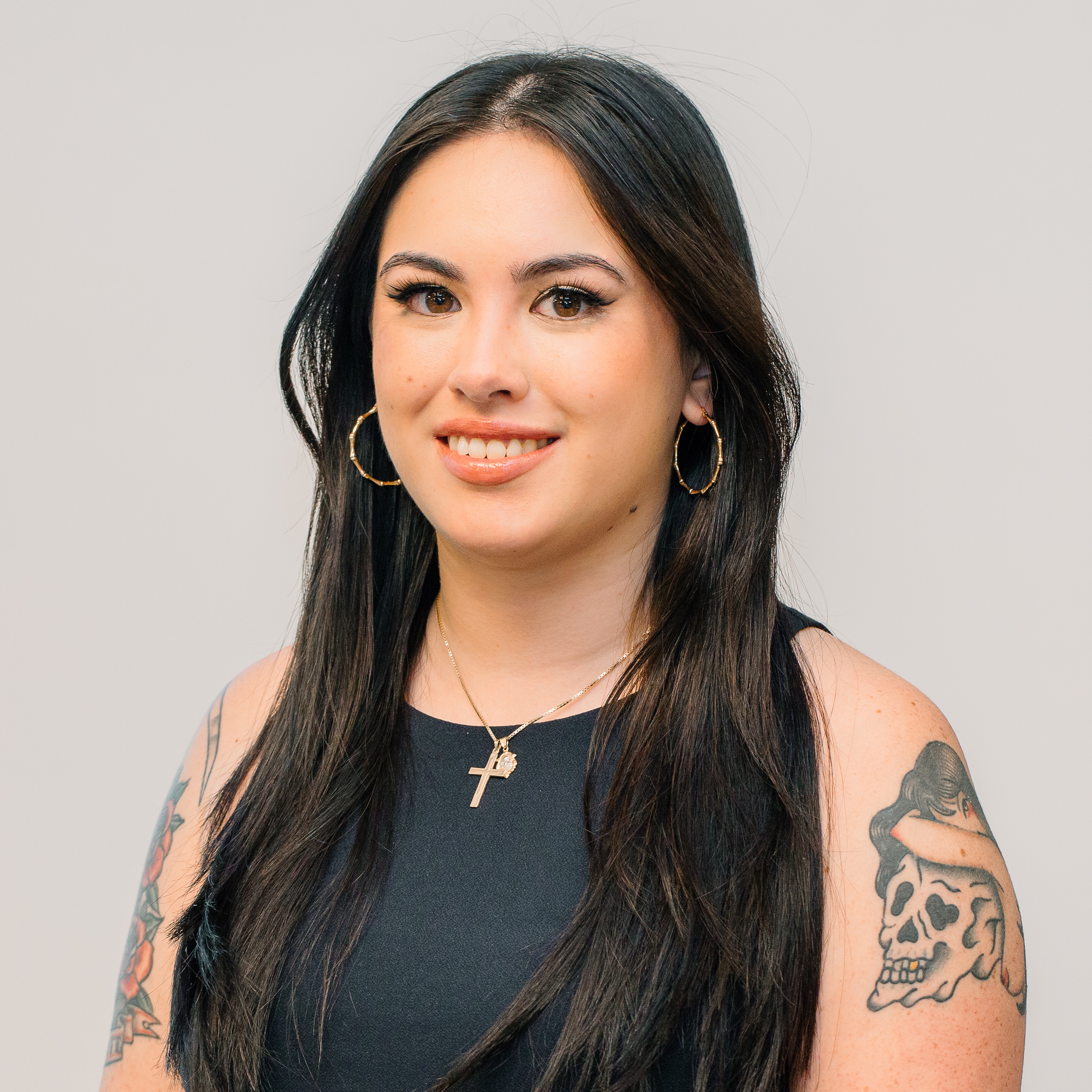 A woman with a black sleeveess shirt and gold hoop earrings posing against an off-white background.