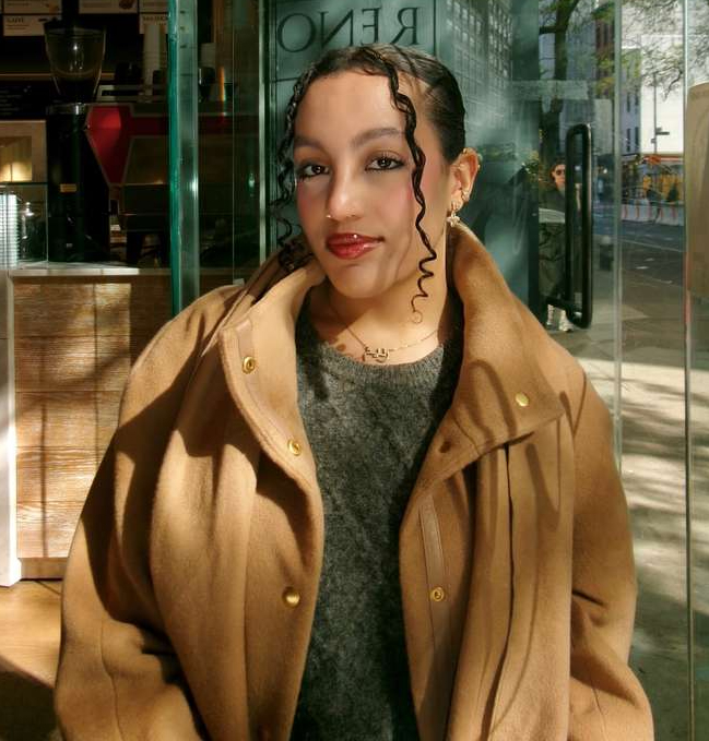 A woman in a beige coat and green sweater standing in dappled sunlight in front of a glass door.