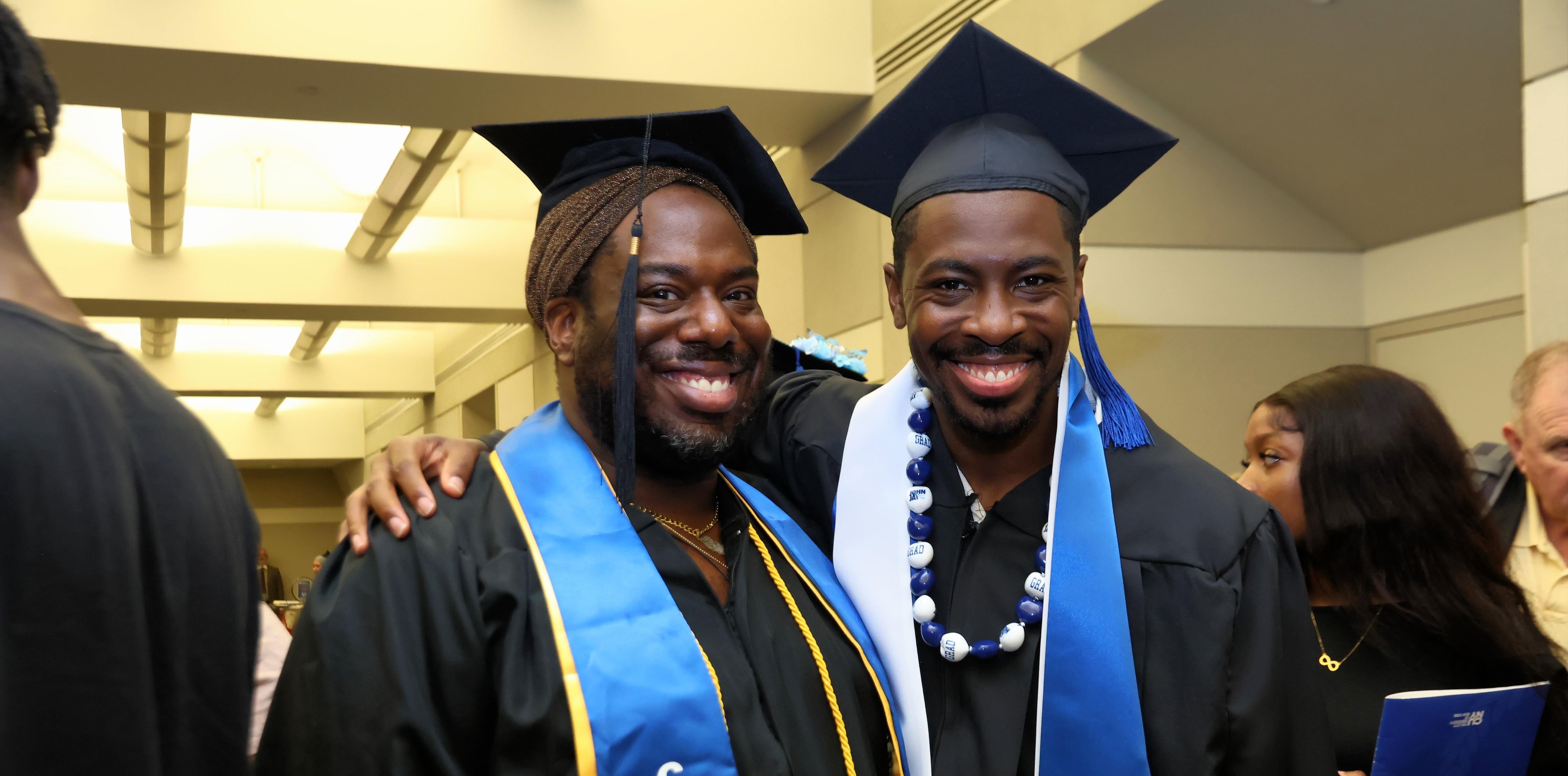 Two CUNY BA graduates in their caps and gowns, one with his hand on the other's shoulders, posing for a photo with big smiles on their faces.