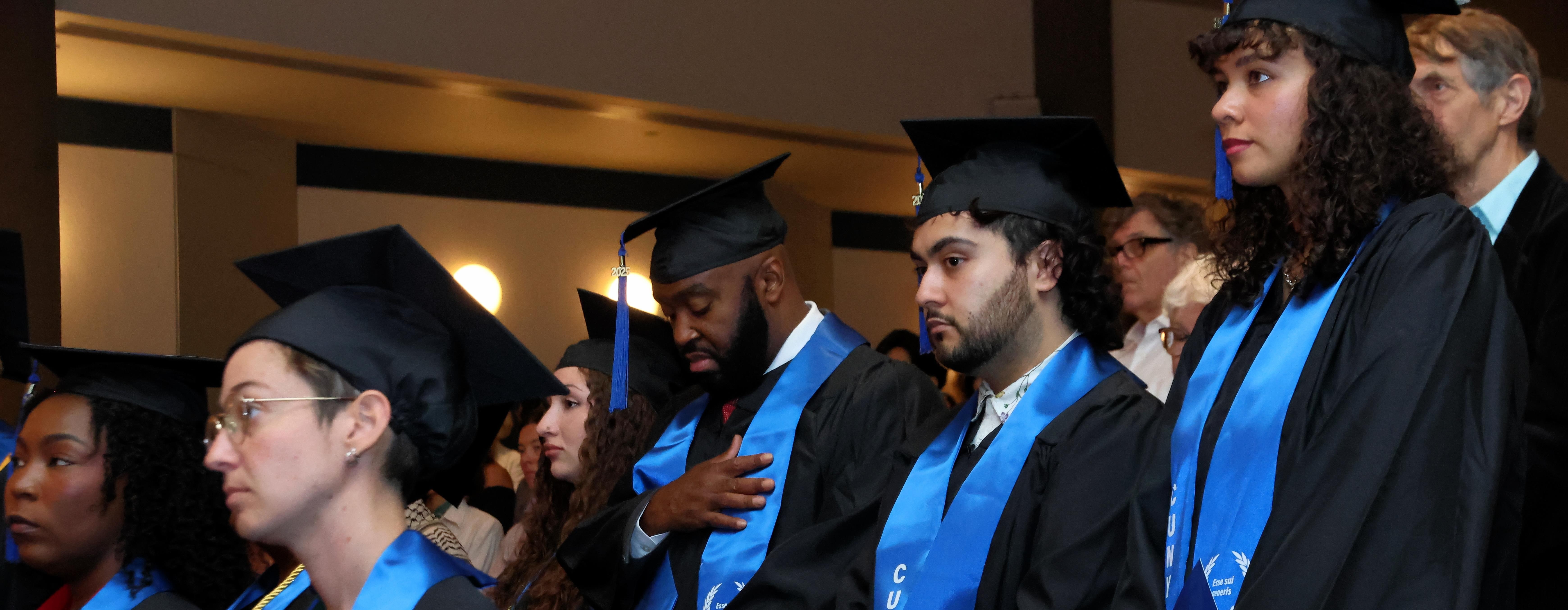 CUNY BA Students dressed in their caps and gowns standing up in front of their seats at their graduation ceremony.