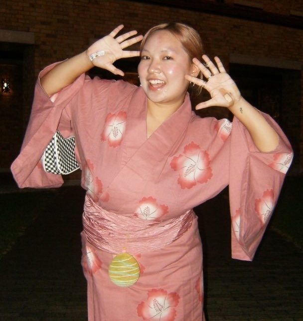 Photo of a woman in a pink kimono posing with her open palms next to her face against a black background.