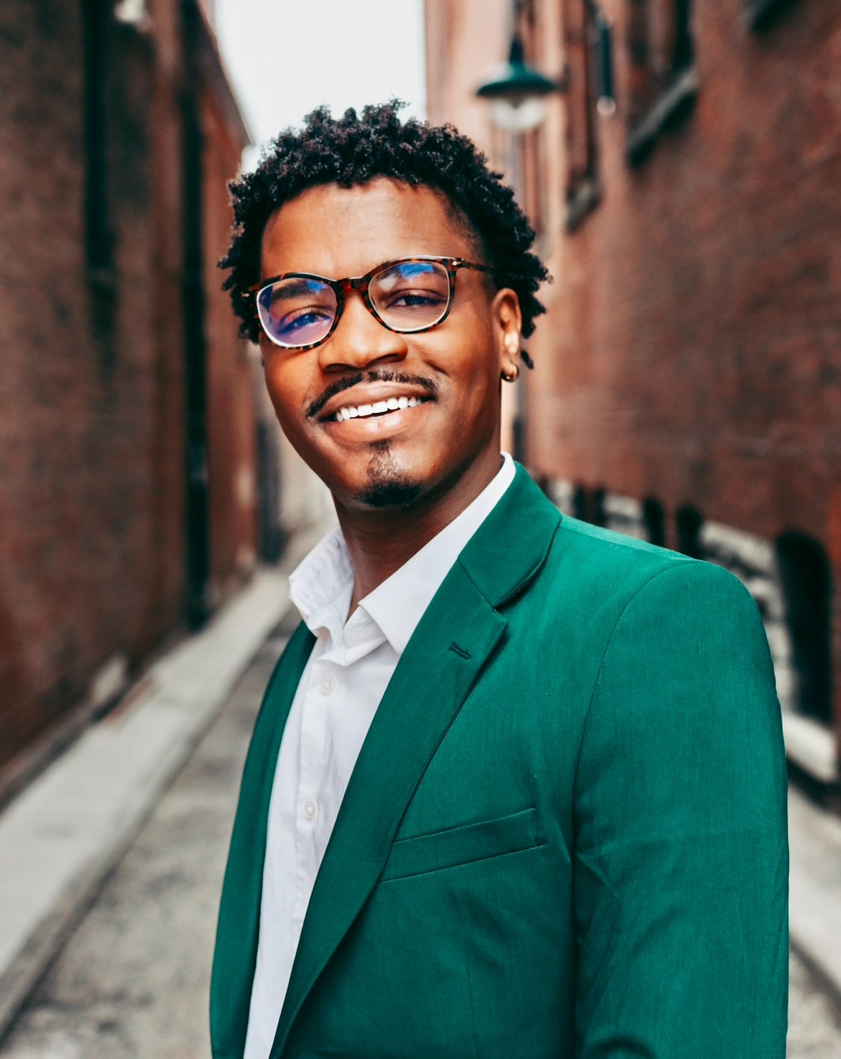 Portrait photo of a man with glasses and a green blazer over a button down shirt posing on a narrow street flanked by tall red-brick buildings on either side.