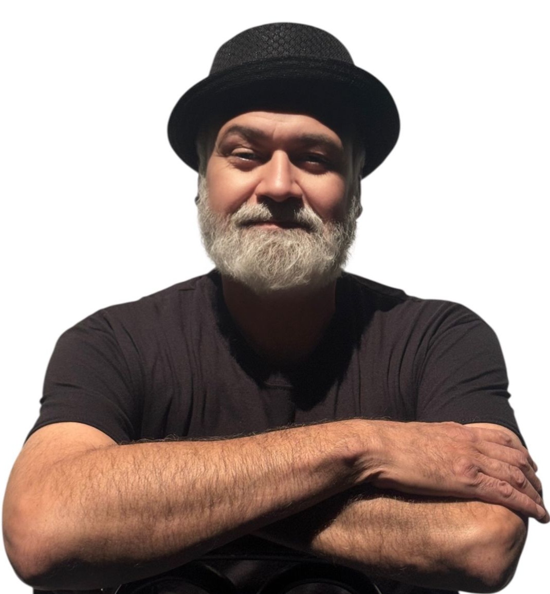 A man in a bowler hat and a black short sleeve t shirt standing with his arms crossed in front of a white background.