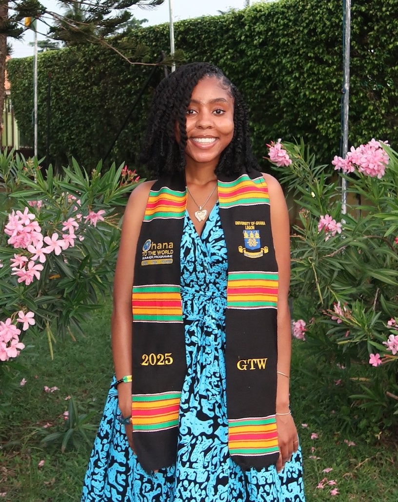 Photo of a woman in a blue dress with a black, green and orange academic stole on her shoulders, standing among pink flowers.