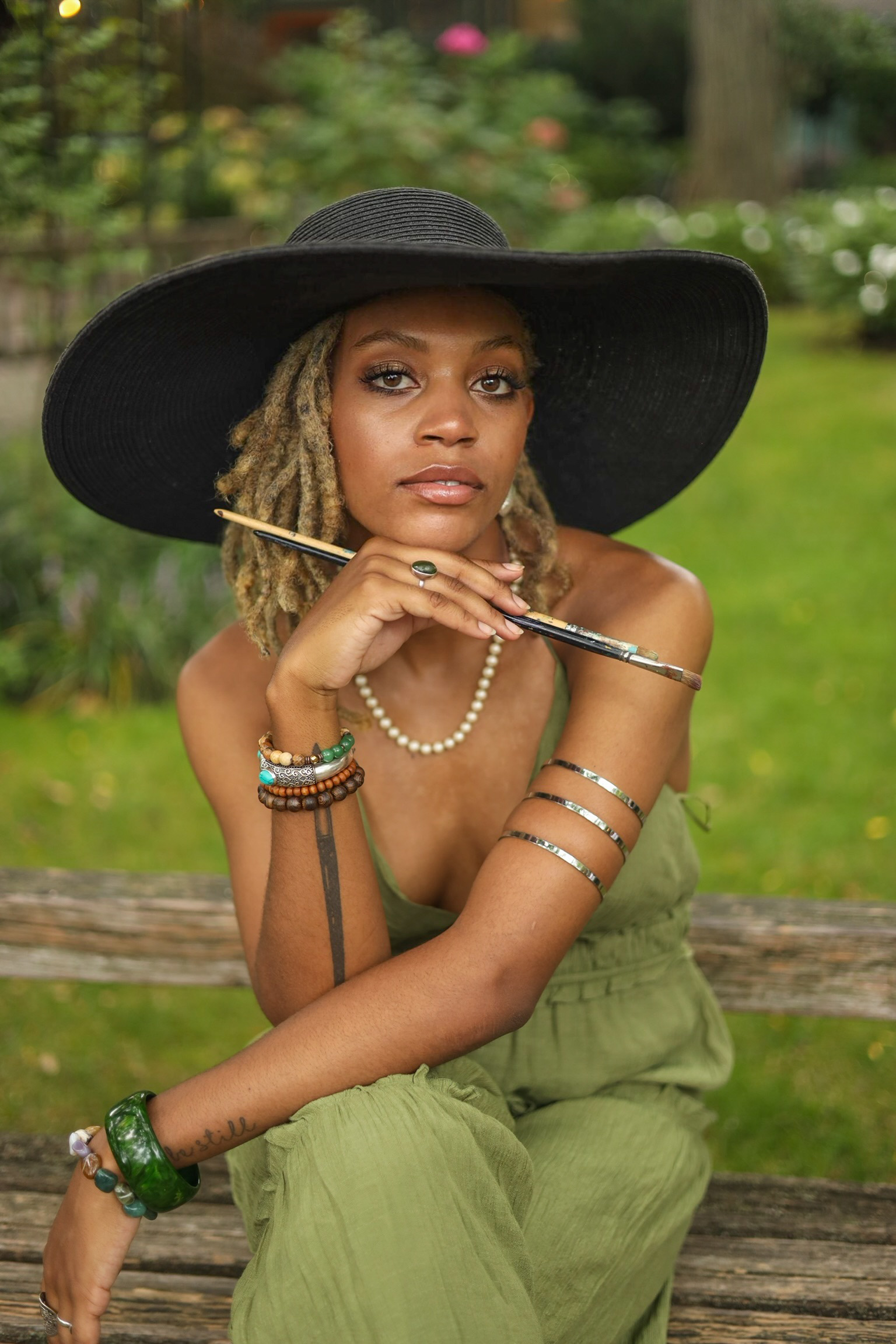 Photo of a woman with a black wide brim hat and a green jumpsuit, posing with two paintbrushes in her upturned right hand.