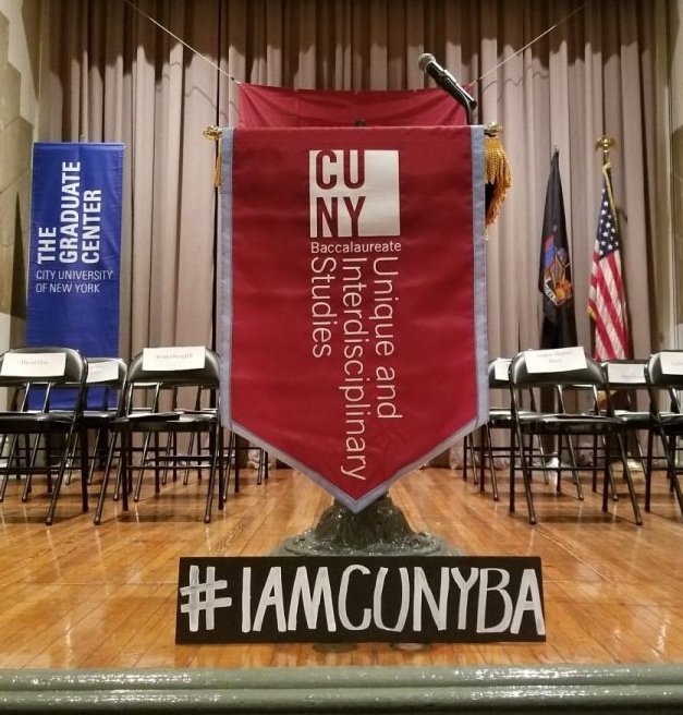 CUNY BA Banner with a sign bearing the hashtag #IAMCUNYBA below it on a stage with a Graduate Center banner to the left and an American flag to the right