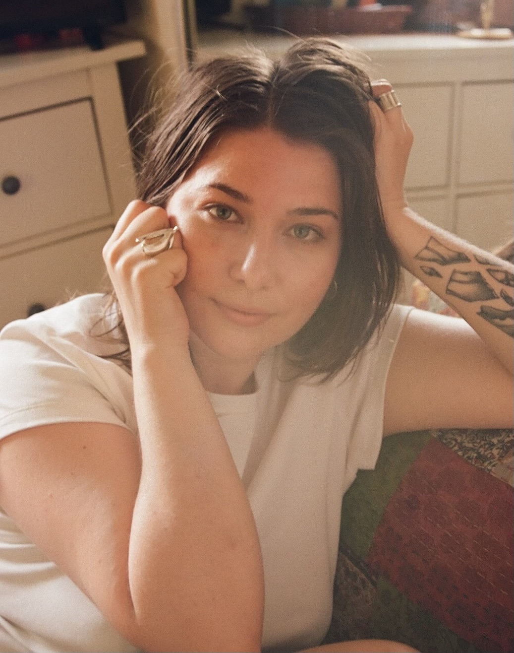 A woman wearing a white shirt, sitting on a couch and leaning her head on her left arm while holding her right hand to the side of her face.